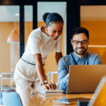 Two colleagues in an office look at a laptop together; one is seated and typing while the other stands beside, both appearing focused and engaged.