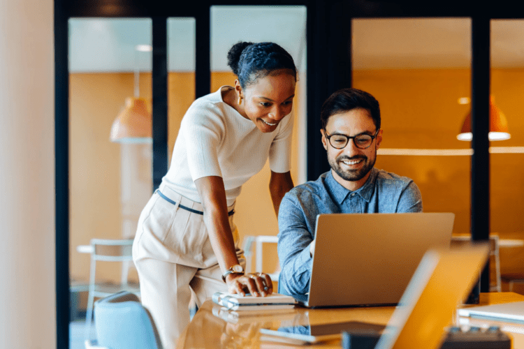 Two colleagues in an office look at a laptop together; one is seated and typing while the other stands beside, both appearing focused and engaged.