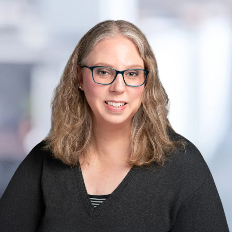 A woman with shoulder-length light brown hair and glasses is wearing a black top and smiling at the camera against a blurred background.