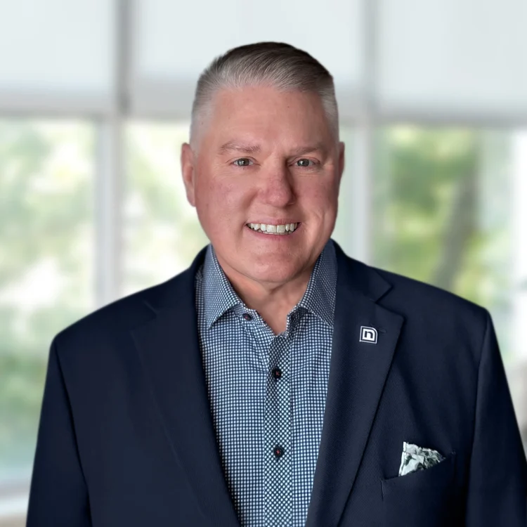 A man in a navy blazer and patterned shirt stands indoors, smiling at the camera with blurred windows and greenery in the background.
