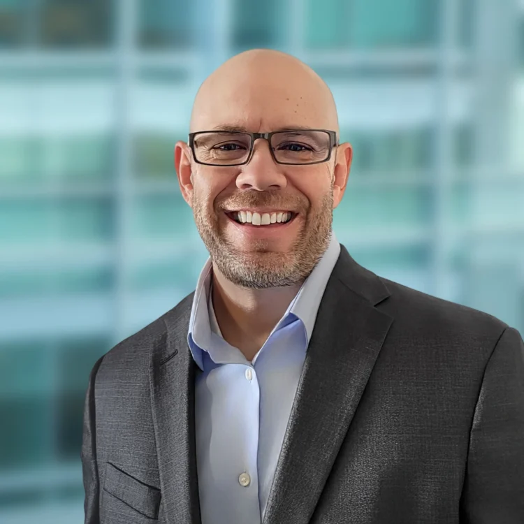 A man wearing glasses and a gray suit jacket over a light blue shirt smiles, standing in front of a blurred glass window background.