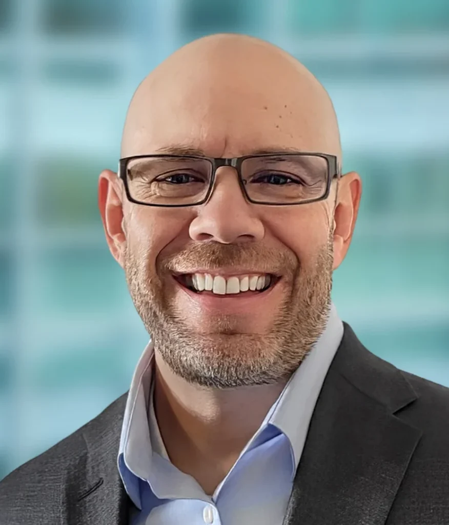 A man wearing glasses and a gray suit jacket over a light blue shirt smiles, standing in front of a blurred glass window background.