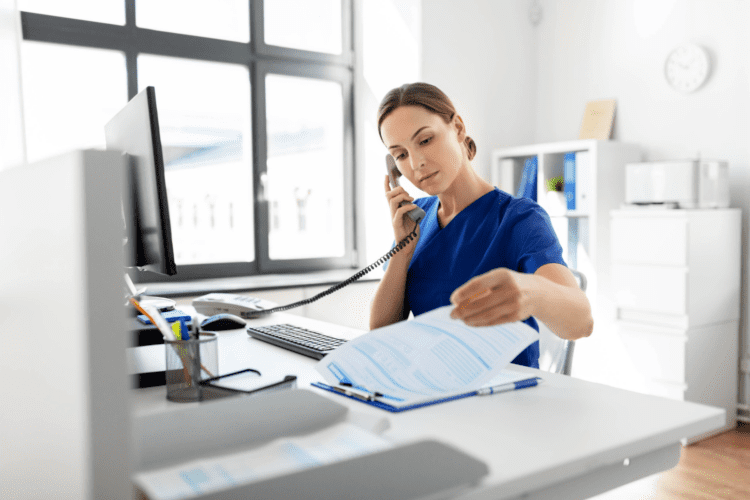 A woman in blue medical scrubs sits at a desk, talking on a phone and reviewing a document in a bright office setting.