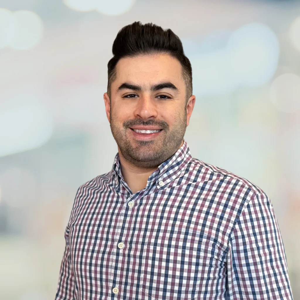 A man with dark hair and a beard wearing a checkered button-up shirt smiles at the camera against a blurred background.