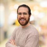 A man with glasses and a beard, wearing a beige knit sweater, stands with arms crossed and smiles at the camera. The background is blurred with warm lighting.