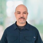 Middle-aged man with a shaved head and short beard, wearing a dark blue collared shirt, standing in front of a blurred light blue and green background.