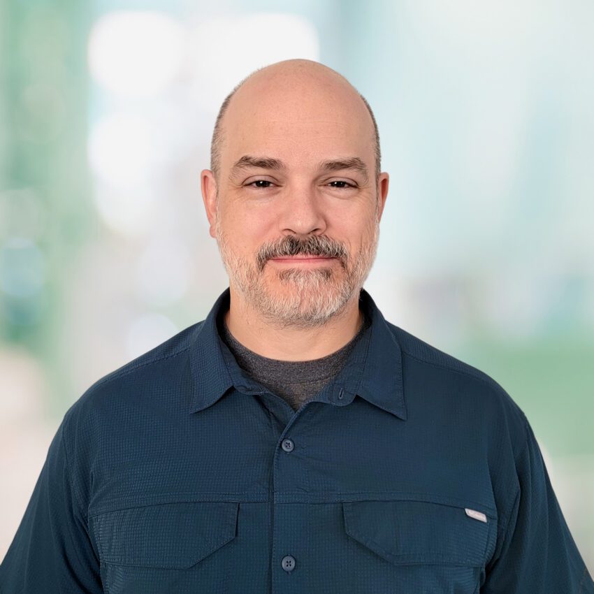 Middle-aged man with a shaved head and short beard, wearing a dark blue collared shirt, standing in front of a blurred light blue and green background.