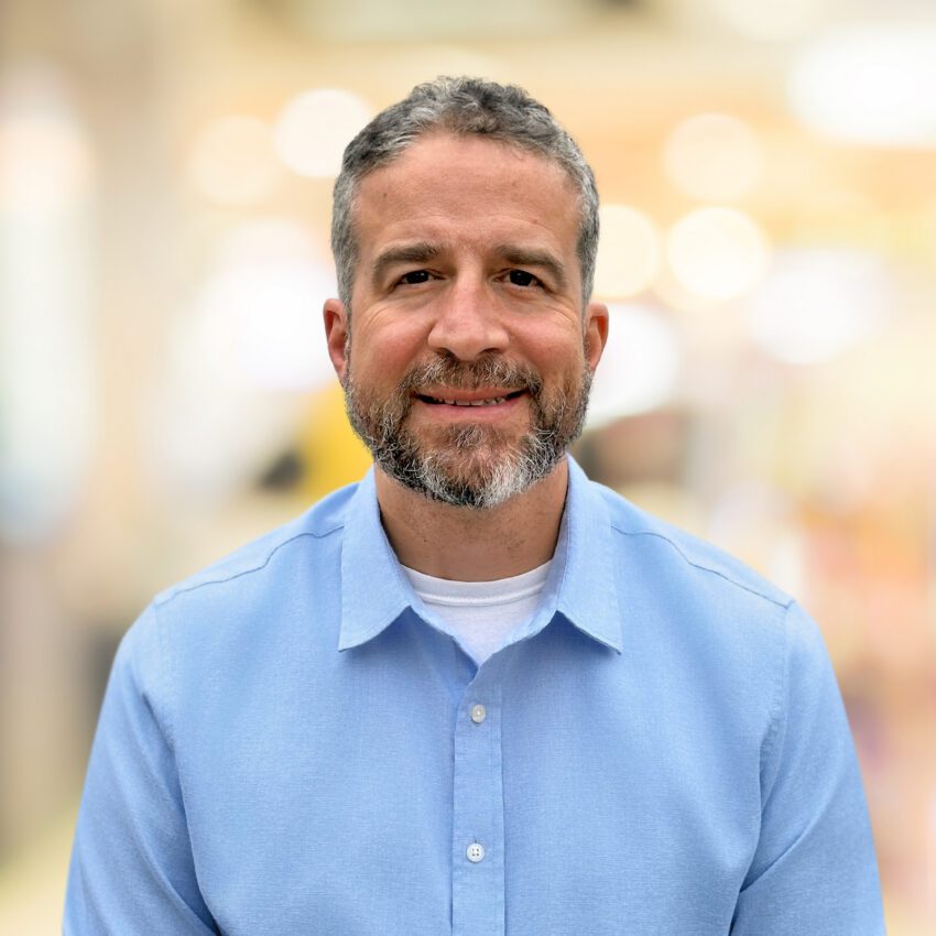 A man with short curly hair and a beard, wearing a light blue button-up shirt and white undershirt, smiles at the camera. The background is softly blurred.