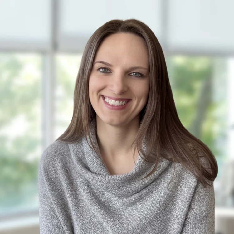 A woman with straight brown hair and a gray sweater smiles while sitting indoors in front of large, blurred windows showing greenery outside.