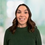 Woman with long brown hair, wearing a dark green sweater, smiles at the camera in a brightly lit indoor setting with a blurred background.