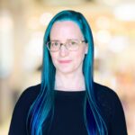 A person with long teal hair, wearing glasses and a black top, stands in front of a softly blurred indoor background.
