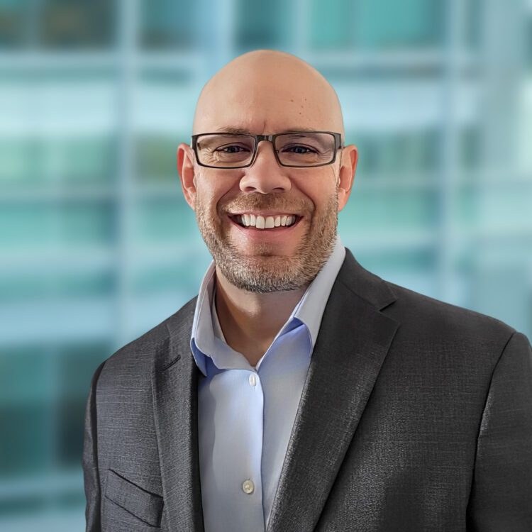 A bald man wearing glasses and a gray suit jacket over a light blue shirt is smiling in front of a blurred office window background.