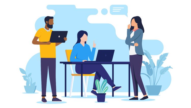 Three people discuss in an office setting; one sits at a desk with a laptop while the other two stand, one holding a laptop. Potted plants decorate the space.