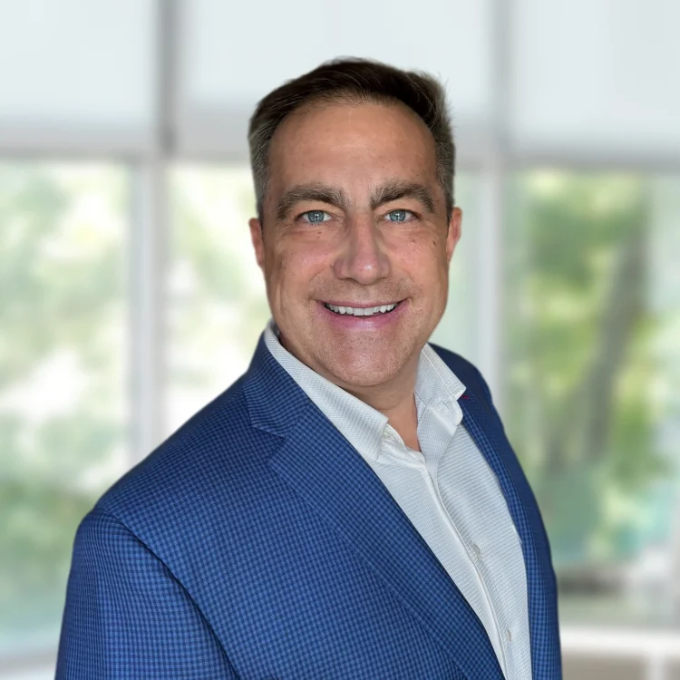 A man in a blue checked blazer and white shirt smiles while standing indoors in front of large windows with a blurred green outdoor background.