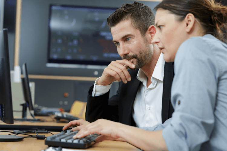 Two people sitting at a desk, looking intently at a computer screen, with one person typing on the keyboard and the other observing closely.