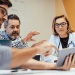 Four professionals, including a doctor, sit at a table in a meeting room discussing healthcare AI governance information displayed on a tablet.