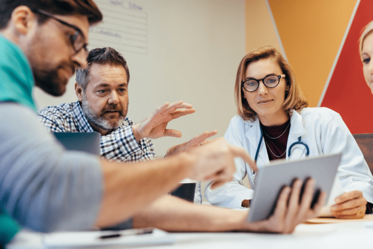 Four professionals, including a doctor, sit at a table in a meeting room discussing healthcare AI governance information displayed on a tablet.