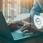 Person typing on a laptop with digital security icons and a shield symbol overlayed, representing cybersecurity and data protection.