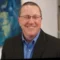 Man wearing glasses, a blue shirt, and a black blazer smiles at the camera; abstract blue artwork and a white wall are visible in the background.